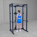 A woman in a blue athletic outfit stands inside a black power rack with a cable pulley system, viewed from behind in a concrete-floored indoor gym space.