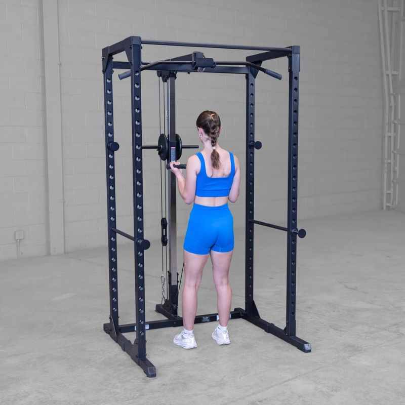 A woman in a blue athletic outfit stands inside a black power rack with a cable pulley system, viewed from behind in a concrete-floored indoor gym space.