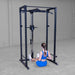 Person sitting on the floor next to a power rack in a room with white walls and gray flooring.
