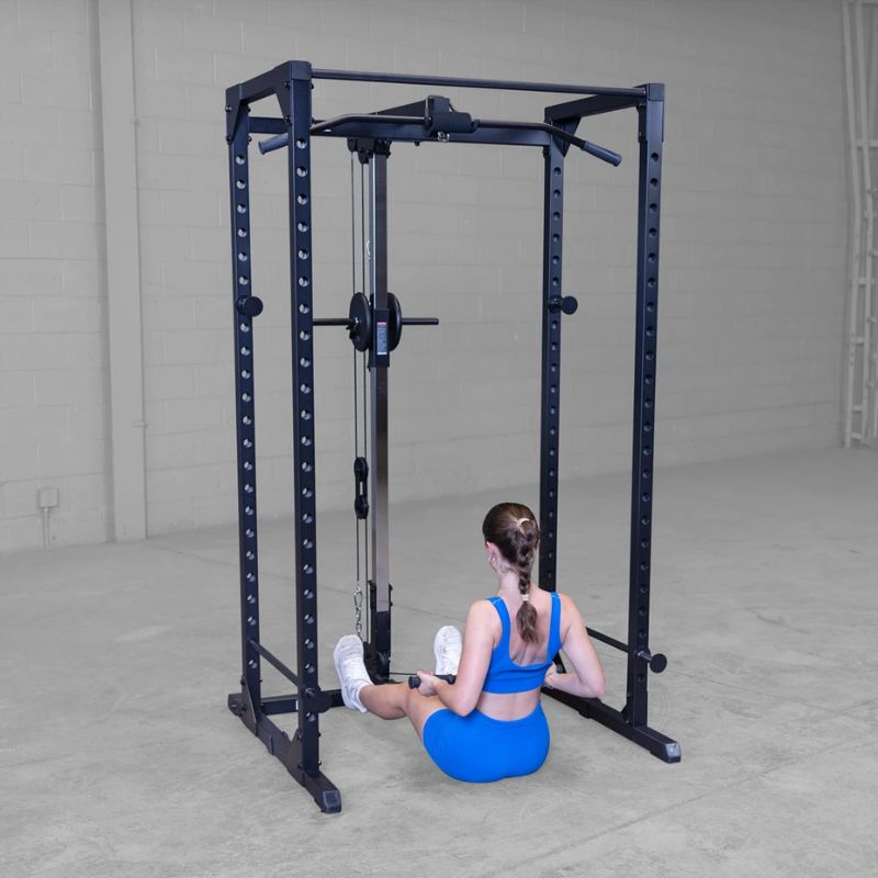 Person sitting on the floor next to a power rack in a room with white walls and gray flooring.