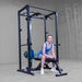 A woman in a blue athletic outfit sits on a weight bench positioned under a black power rack with a barbell loaded with blue, yellow, and green weights, set in a gray indoor gym space.