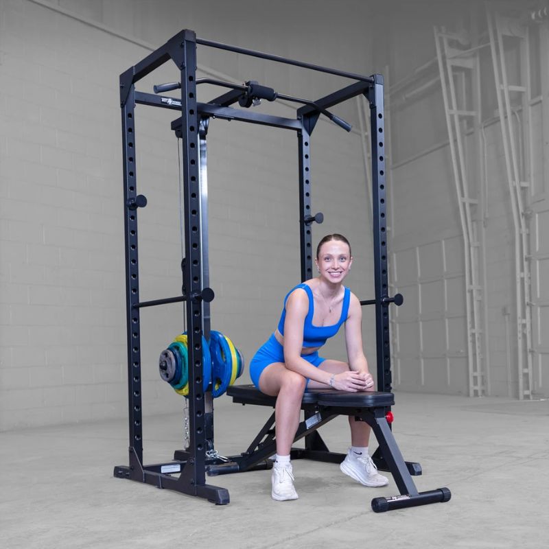 A woman in a blue athletic outfit sits on a weight bench positioned under a black power rack with a barbell loaded with blue, yellow, and green weights, set in a gray indoor gym space.
