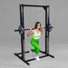 A woman in a green tie-dye crop top and bright green leggings performs a front split squat on a black power rack with a barbell loaded with weight plates, set against a gray concrete wall and floor.