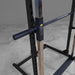 Close-up of a black adjustable power rack with a vertical metal upright, safety stopper, and attached horizontal bar, shown in a gym setting on a concrete floor.