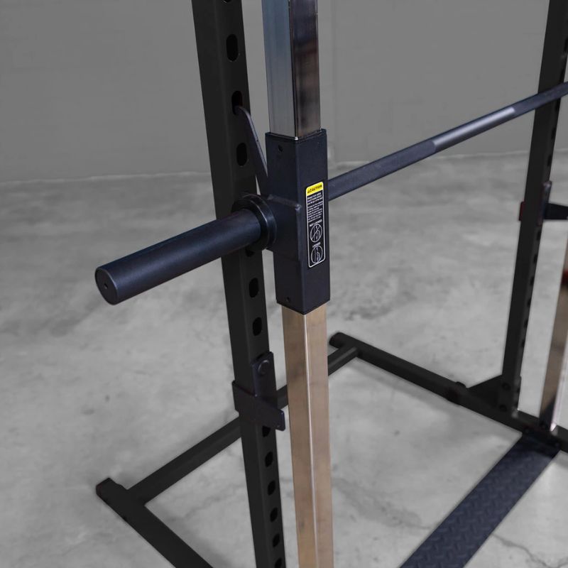 Close-up of a black adjustable power rack with a vertical metal upright, safety stopper, and attached horizontal bar, shown in a gym setting on a concrete floor.